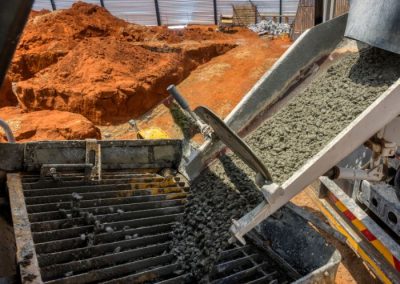 Concrete is being poured from a truck chute into a metal grate at a construction site with red dirt and materials around.