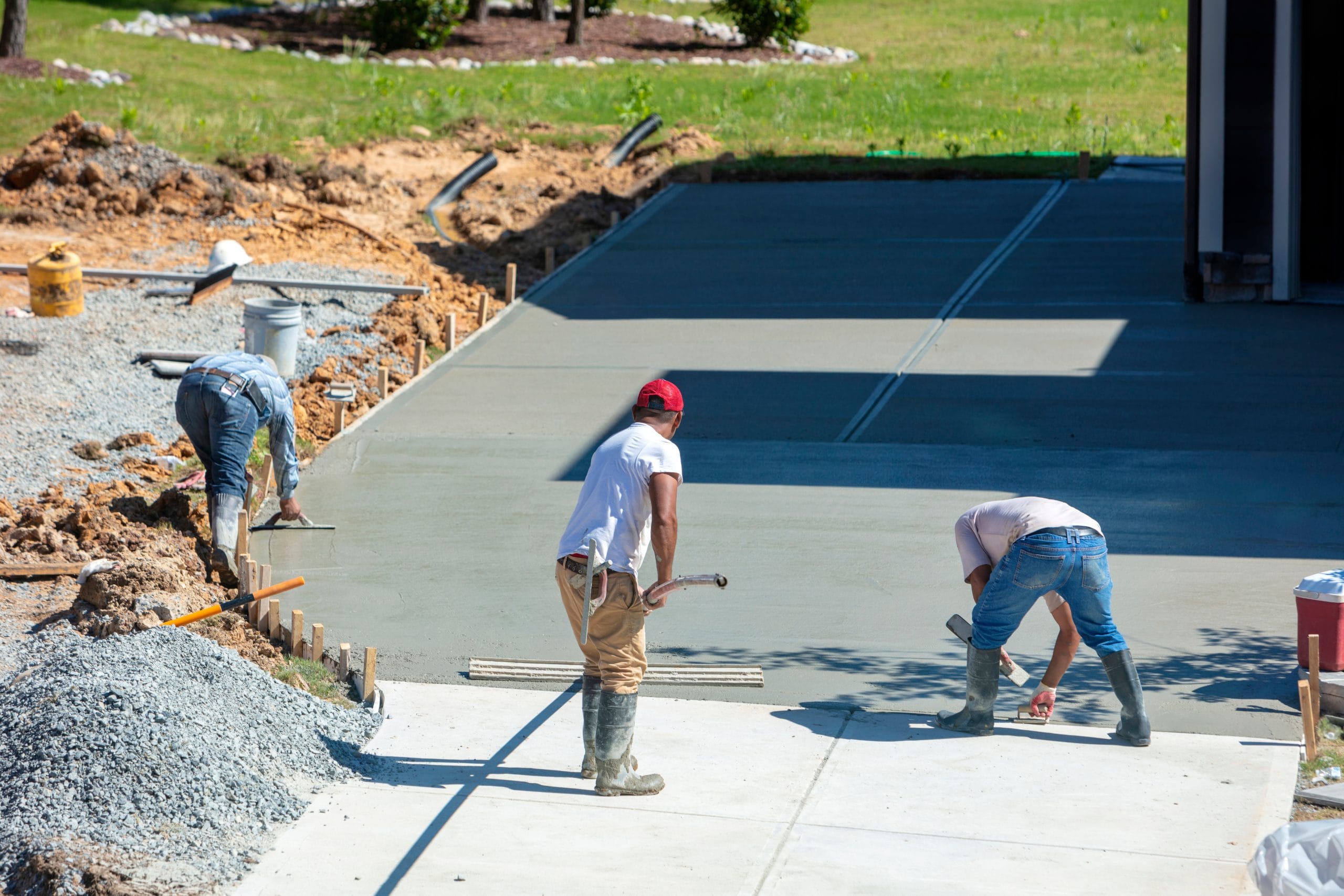 Three workers in boots leveling and smoothing wet concrete on a driveway construction site outdoors.