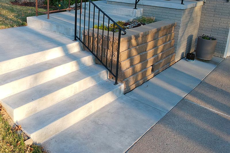 Concrete steps with a black railing and a short brick wall next to a sidewalk in sunlight.