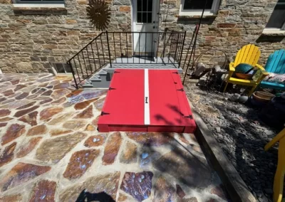 Red storm cellar doors with a white stripe are installed on a stone patio near steps and colorful chairs.