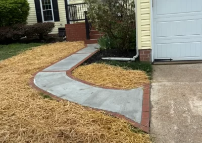 A new concrete walkway bordered with bricks curves from driveway to front steps, with straw covering the yard.