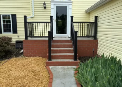 Brick steps with black railings lead to a white door on a yellow house, with plants and straw-covered ground nearby.