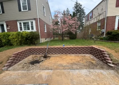 A newly built stone retaining wall frames a bare patch of dirt between two houses with a blooming tree in the background.