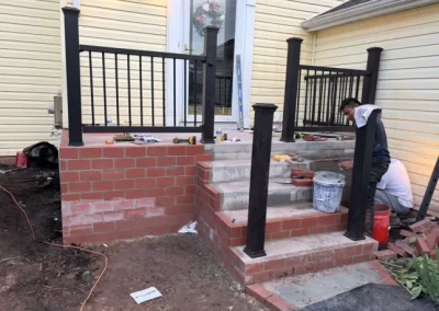 A man builds a brick porch and stairs outside a yellow house with black railings.