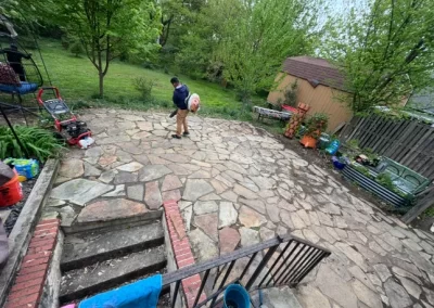 A person walks on a stone patio in a backyard with garden tools, plants, and a shed in view.