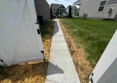 Open white gate revealing a concrete walkway through a grassy yard toward houses in the background.