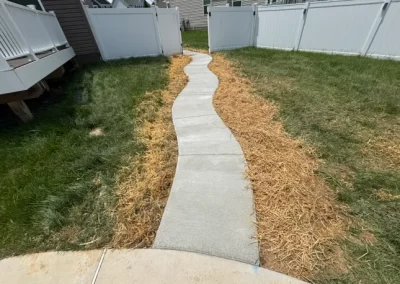A curvy concrete walkway surrounded by straw and green grass in a fenced backyard.