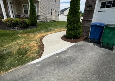 A new concrete walkway curves from a driveway toward a house between green grass and shrubs.