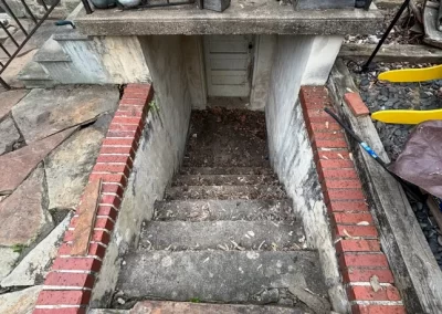 Outdoor stone and brick stairs lead down to a basement door, surrounded by metal railings and scattered tools.