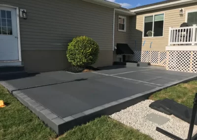 Freshly poured concrete patio beside a beige house, bordered by a small shrub and lattice skirting.