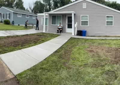 A small gray house with a new sidewalk, people standing and sitting outside, and patches of grass in the yard.