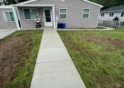 A person sits on the porch of a gray house with a new concrete walkway and freshly patched lawn.