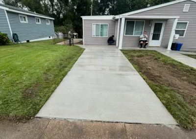 A small gray house with a fresh concrete driveway, green lawn, and a person sitting on the porch.