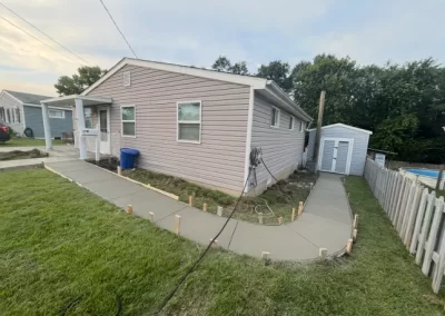 Freshly poured concrete sidewalk curves around a light purple house with a small shed in the fenced backyard.