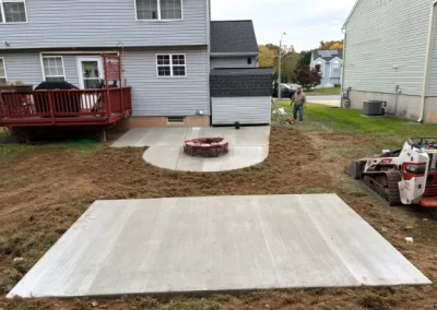 Fresh concrete patio slab and curved walkway with fire pit in backyard of a suburban house, people working nearby.