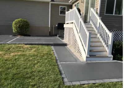 Gray concrete walkway and steps with white railing next to a house, bordered by green grass and a small bush.