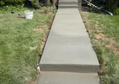 Freshly poured concrete sidewalk with steps leading up to a house entrance, surrounded by grass and bushes.