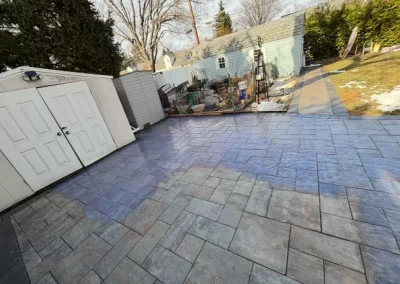 Backyard with a shed, garden, and a large area of patterned concrete pavers in sunlight.