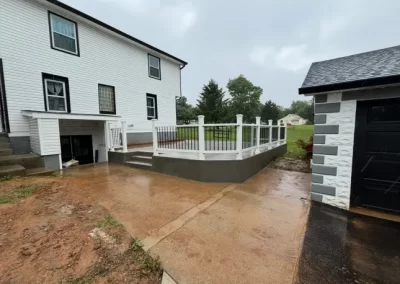 A concrete ramp with white railings leads to a porch beside a white house on a rainy day.