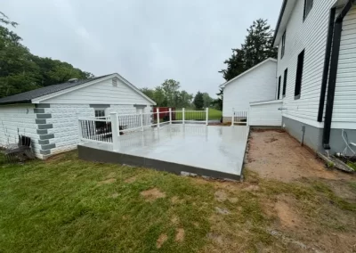 A new concrete patio with white railings next to a white house, bordered by grass and dirt.