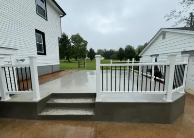 A small concrete patio with white railings and steps, next to a white house and green lawn on a cloudy day.