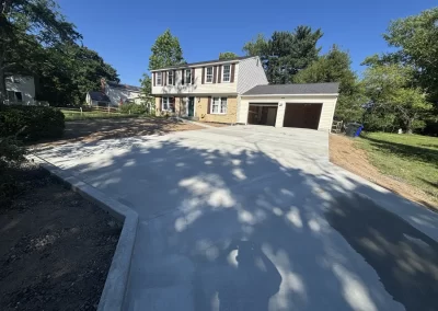 Two-story suburban house with a large, freshly paved concrete driveway under a clear blue sky.