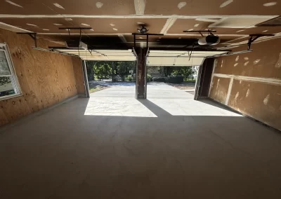 Empty two-car garage with open doors, unpainted drywall, and sunlight shining onto the concrete floor.