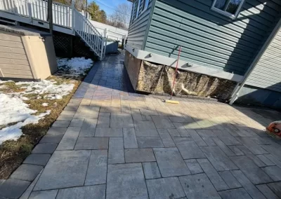 A broom rests on a sunlit patio with stone pavers beside a house; some snow remains on the grass.