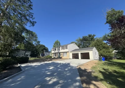 Two-story suburban house with a new concrete driveway and double garage, surrounded by trees on a sunny day.