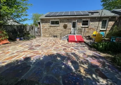 Stone patio in backyard, two chairs, dog ramp, and solar panels on roof, under clear blue sky.