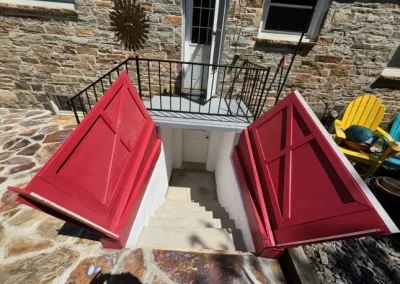Open red cellar doors reveal stairs leading down to a basement entrance of a stone house.