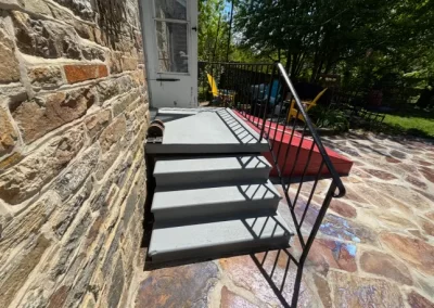 Gray steps with a black railing lead to a white door, beside a stone wall and a sunlit patio with chairs.